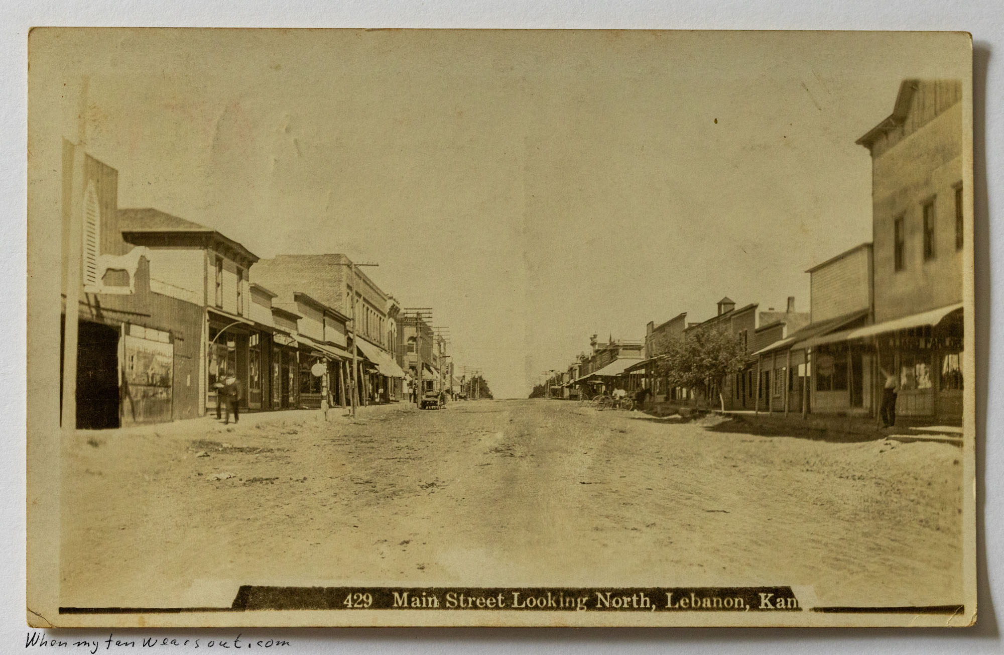 Sepia-toned historical postcard showing a wide, unpaved Main Street in Lebanon, Kansas, looking north. The street is lined with two-story commercial buildings featuring awnings, and a few indistinct figures walk along the dirt street and sidewalks under a bright sky. The caption