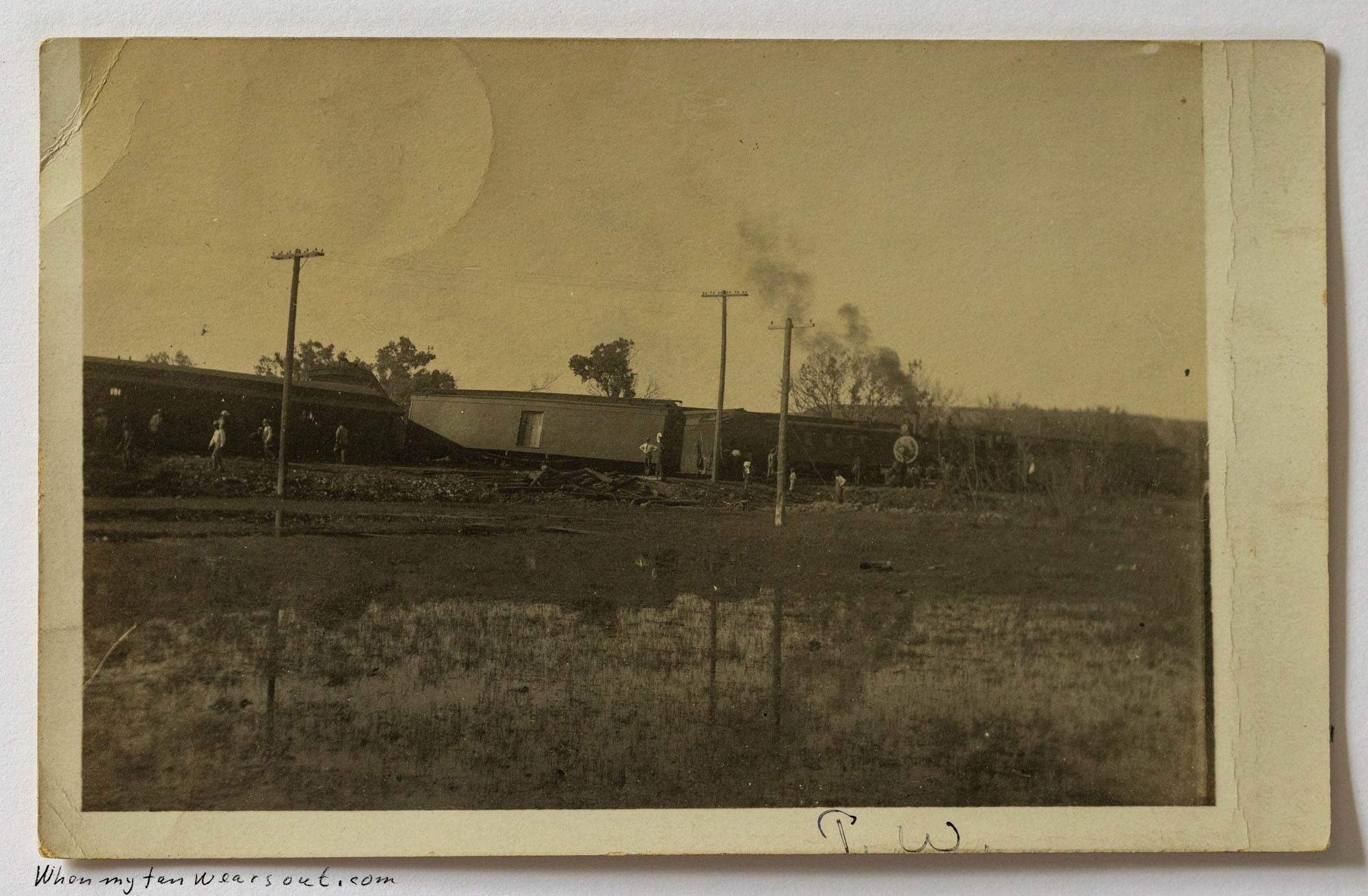 Sepia-toned photograph of a train derailment showing several passenger cars or freight cars overturned or off the tracks, with utility poles stretching across the horizon, a few people inspecting the damage, and a flooded or muddy field in the foreground, with the faint initials