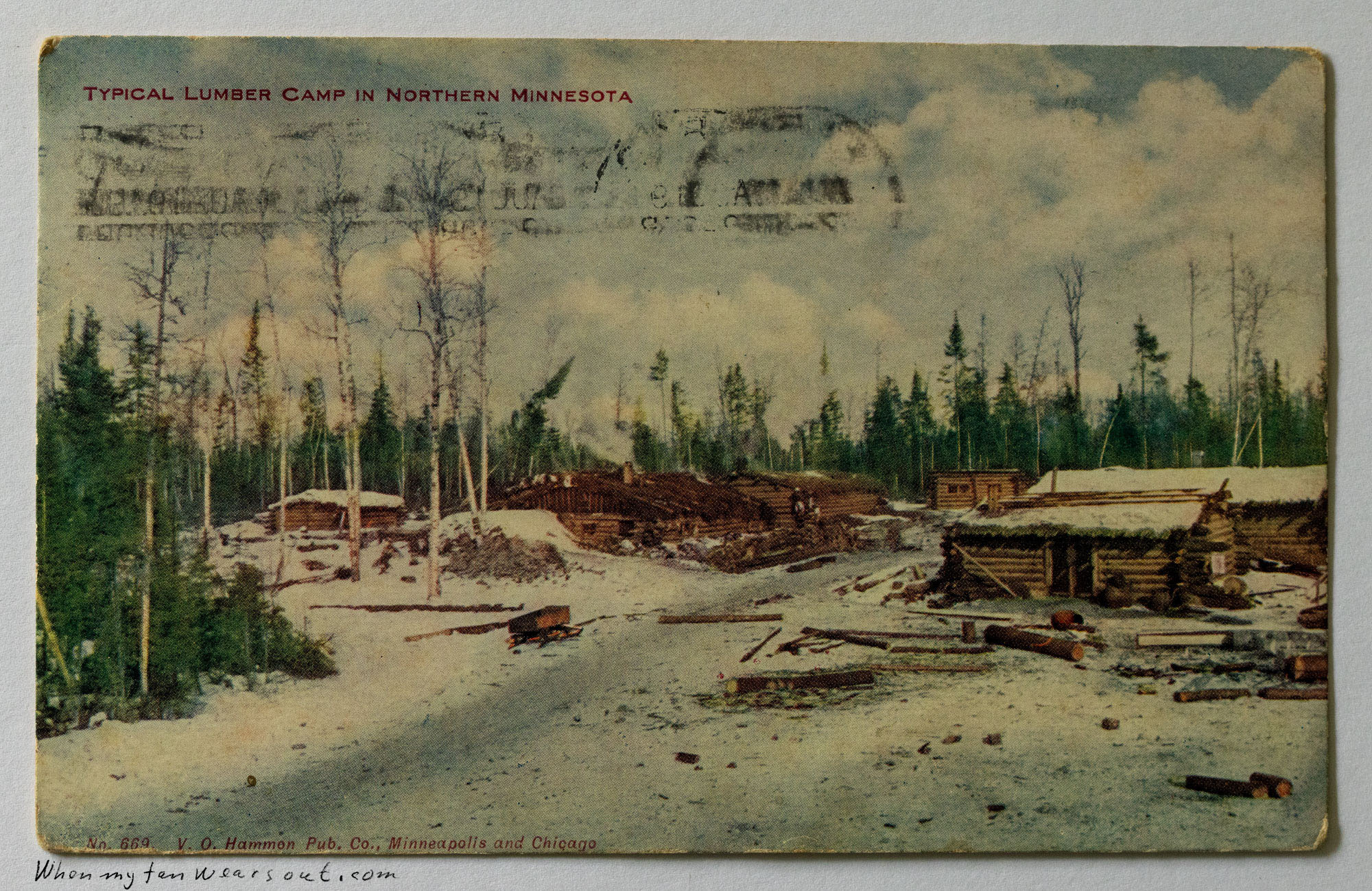 Hand-colored postcard depicting a snowy lumber camp in Northern Minnesota with log cabins, stacked timber, and bare trees under a cloudy sky, titled