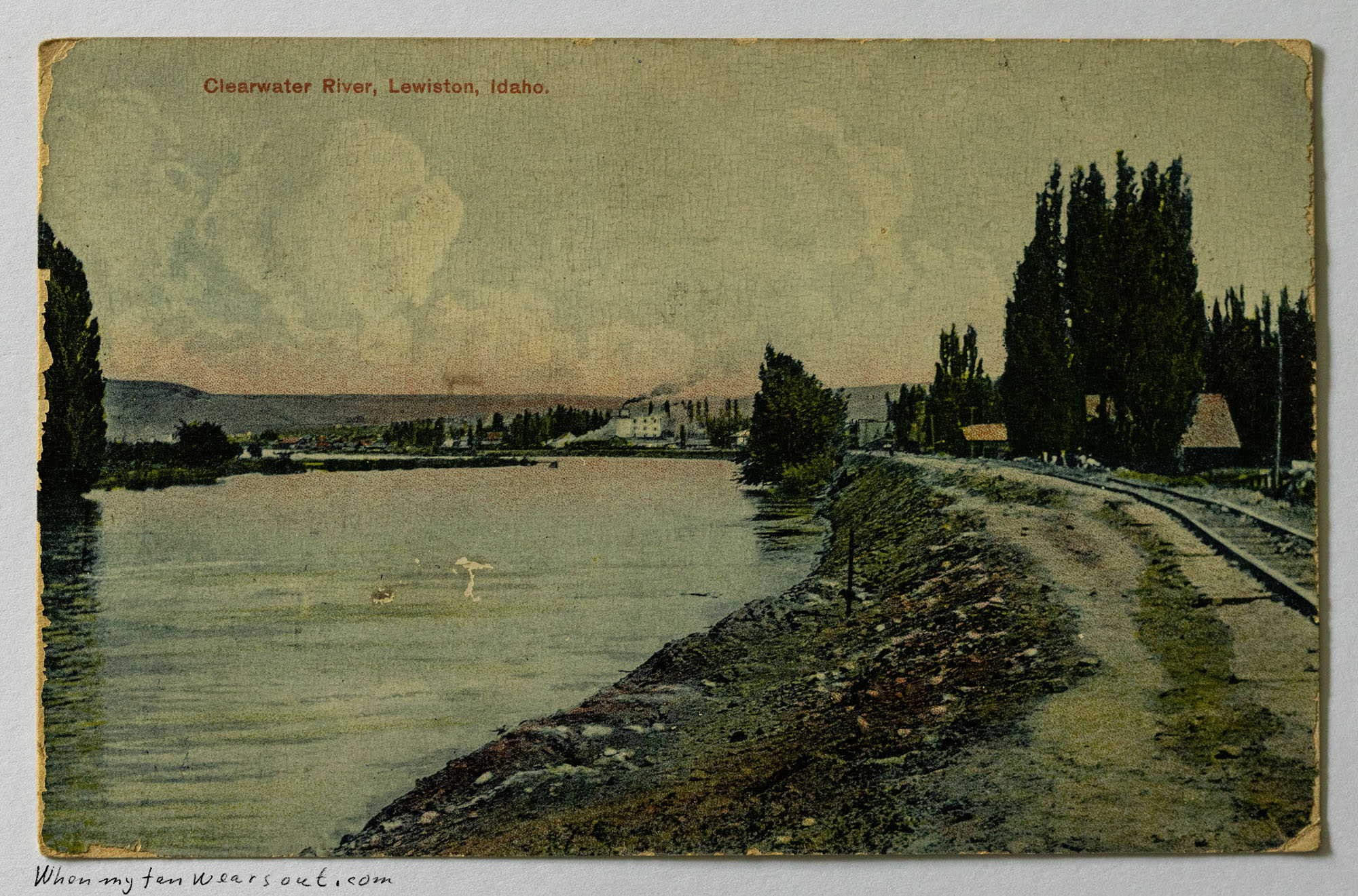 A vintage postcard depicting the Clearwater River in Lewiston, Idaho. The river dominates the foreground with calm, reflective water. Along the right bank, a raised railroad track runs parallel to the river, with tall, slender trees lining its side. In the background, across the river, lies the town of Lewiston nestled against distant hills under a cloudy sky. The sky above is a mix of pale blues and whites, with a hint of warm colors from the postcard's printing process. The title
