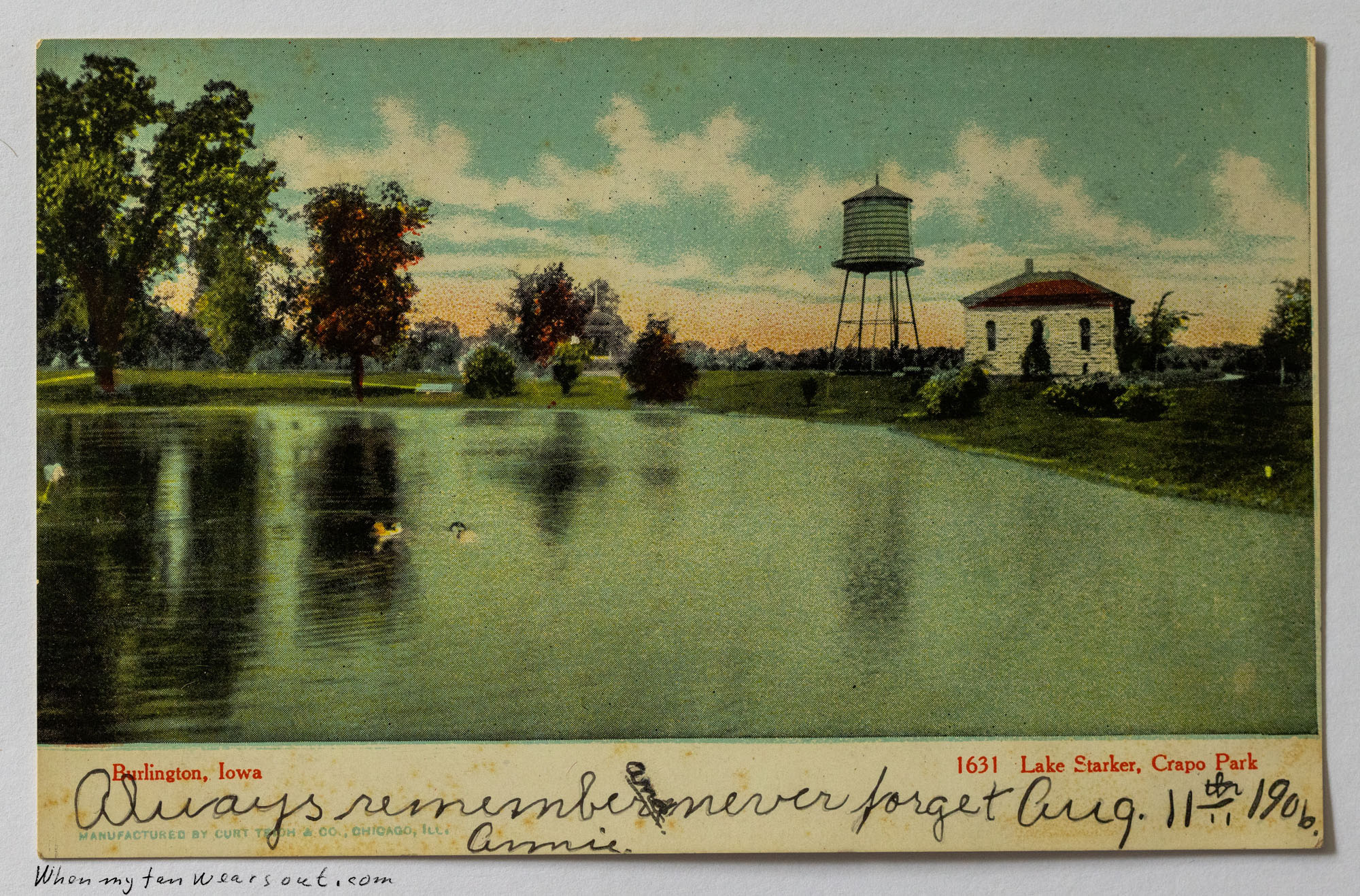A hand-colored vintage postcard depicts a serene green lake reflecting trees on the left, with two ducks on the water. On the right bank, a tall metal water tower stands next to a white building with a red roof under a light blue sky with white clouds.