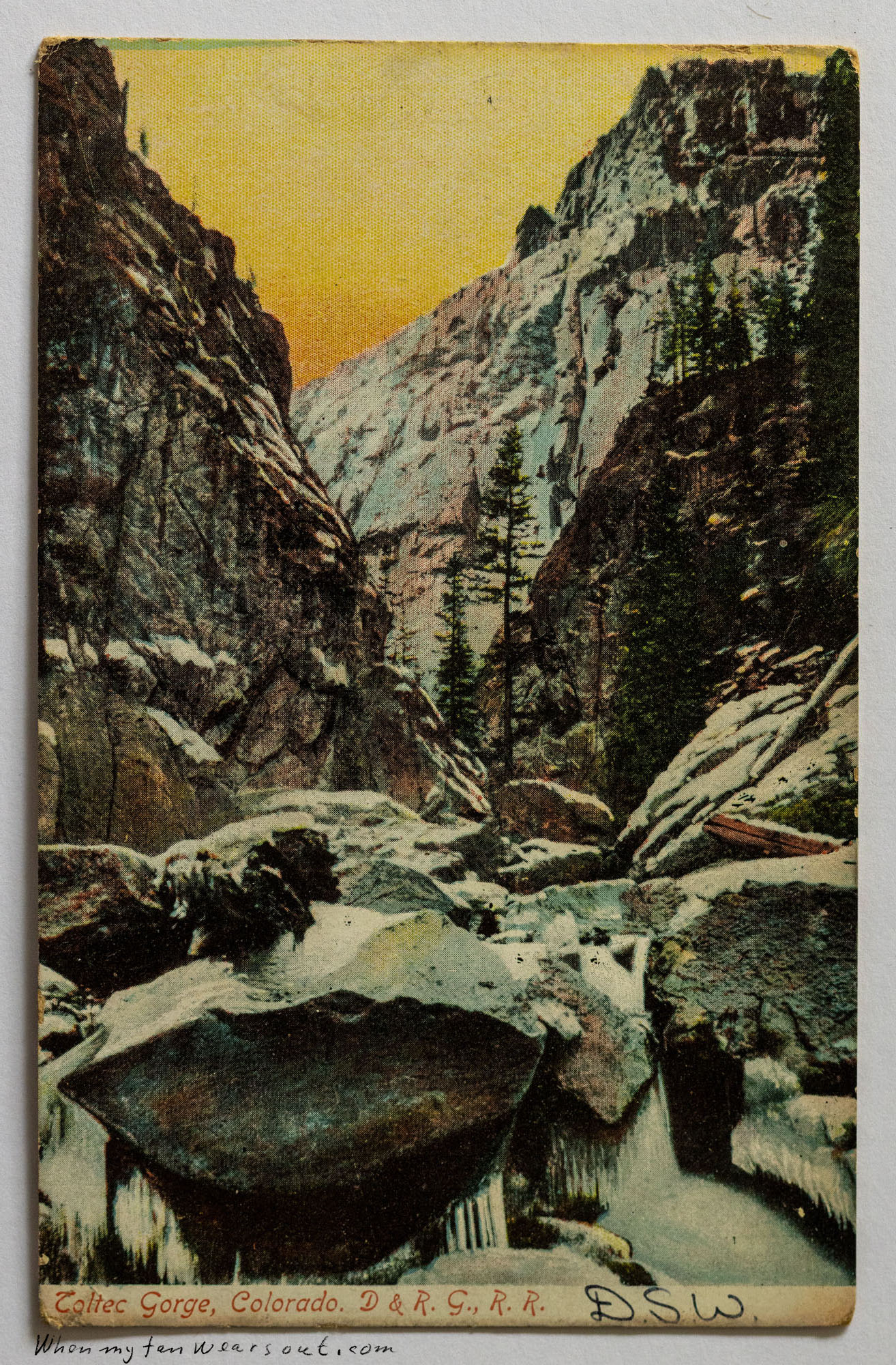 Vintage postcard showing a hand-colored image of the Toltec Gorge in Colorado, with a river flowing through large boulders, snow-covered banks, and steep rocky cliffs under a golden, orange-hued sky, with scattered evergreen trees.