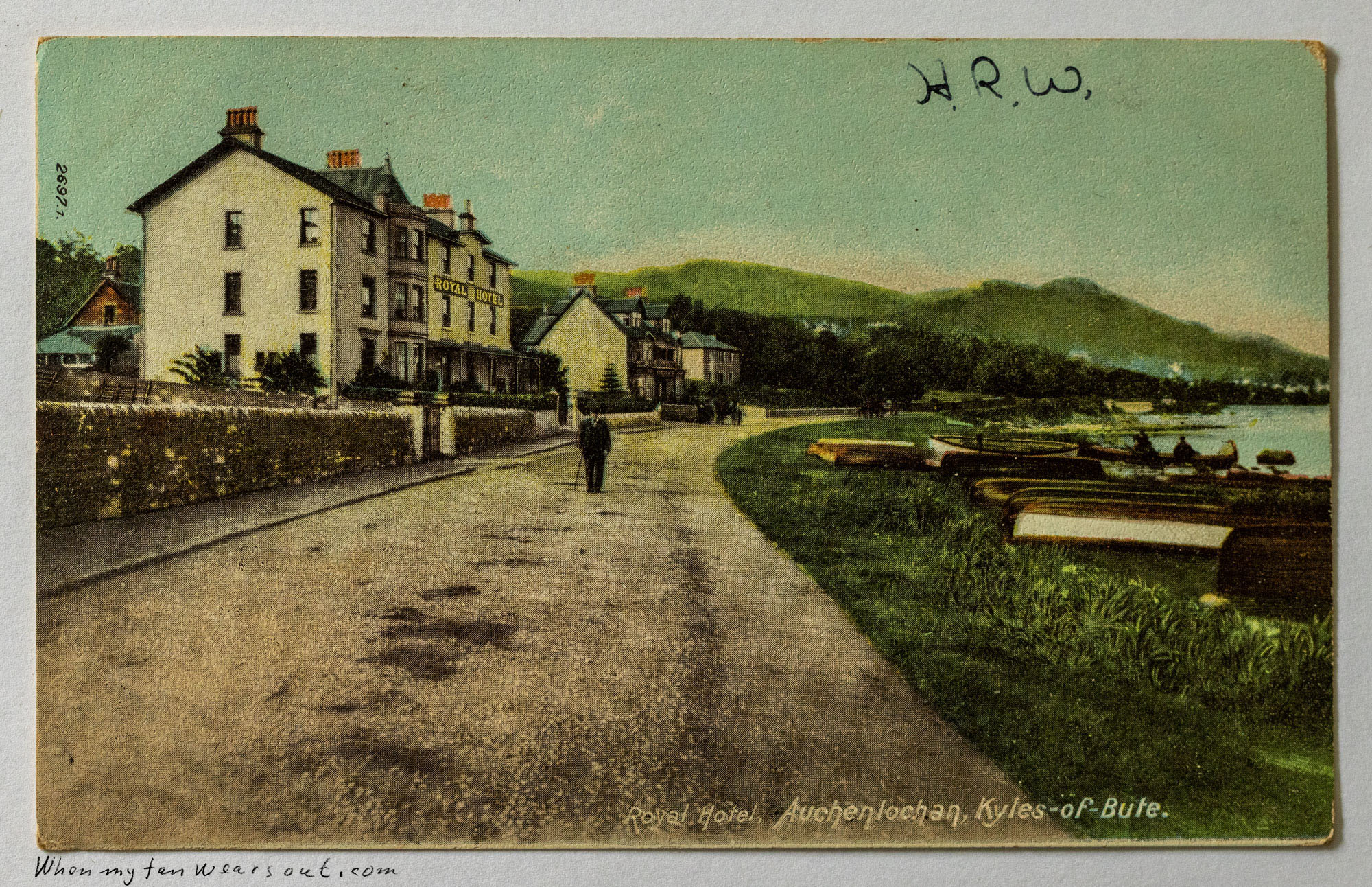 A vintage hand-tinted postcard featuring a scenic view of the Royal Hotel in Auchterlochan, Kyles-of-Bute, Scotland. The image shows a large white hotel building next to a paved road, where a lone male figure walks. To the right, several small boats are moored in a calm body of water, with rolling green hills in the distance under a light blue sky. Handwritten initials