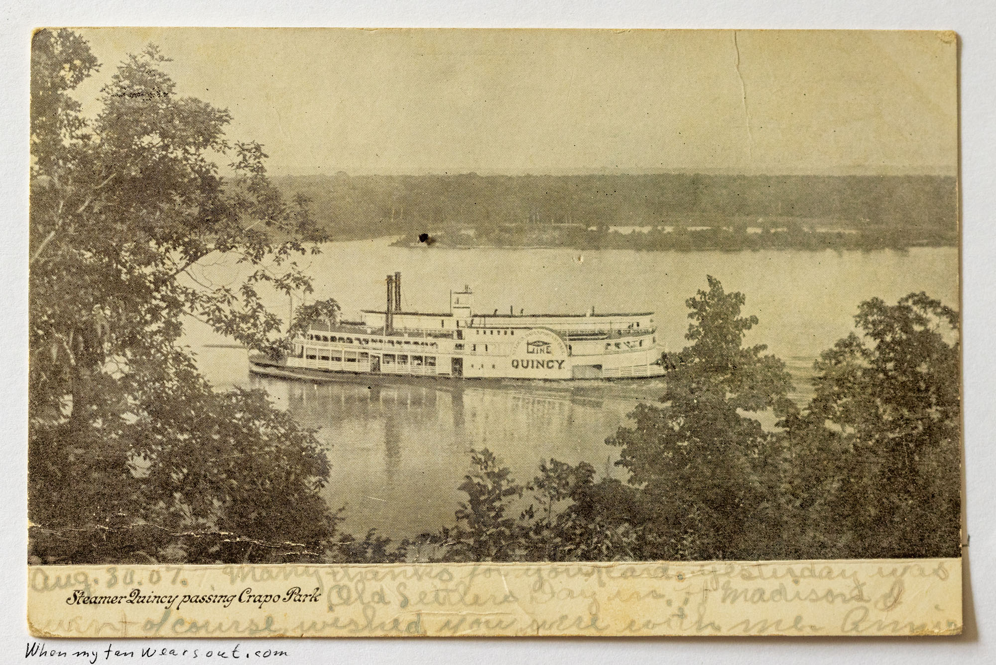 Front of postcard #0066: Sepia-toned photograph of the paddle-wheel steamboat "Quincy" on a river, viewed from an elevated bank through framing t