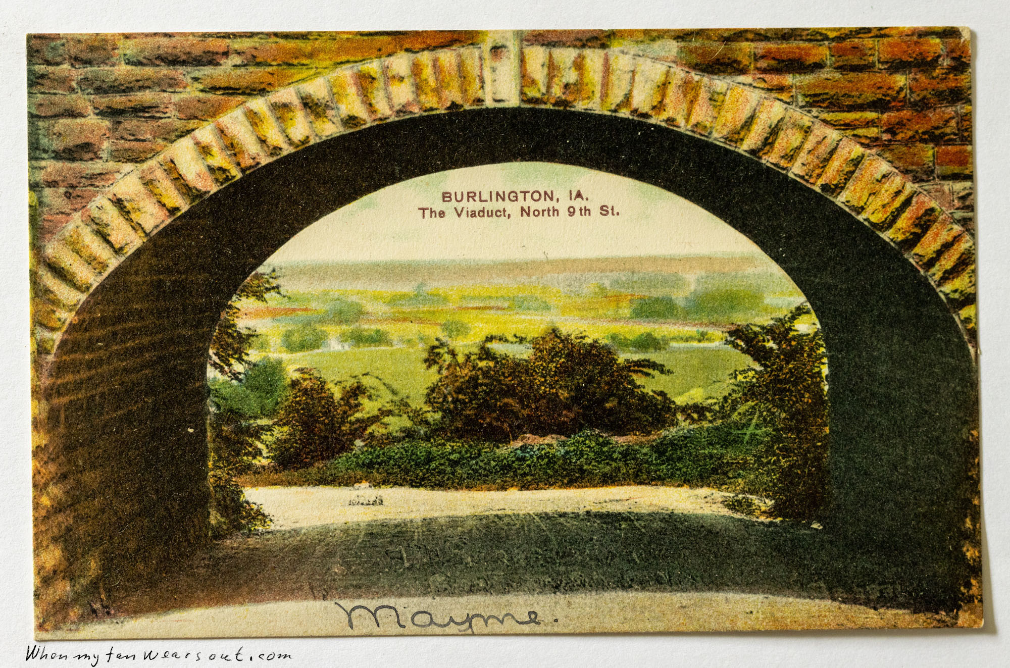 Front of postcard #0082: Color illustration of a scenic view looking through a dark brick-arch viaduct. The view frames a landscape of lush green