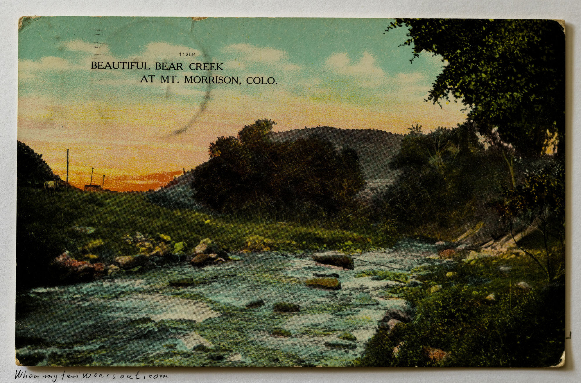 A vintage, hand-colored postcard depicting a serene sunset over Bear Creek at Mount Morrison, Colorado, with water flowing over rocks surrounded by dense green trees and distant hills under a vibrant orange and green sky.