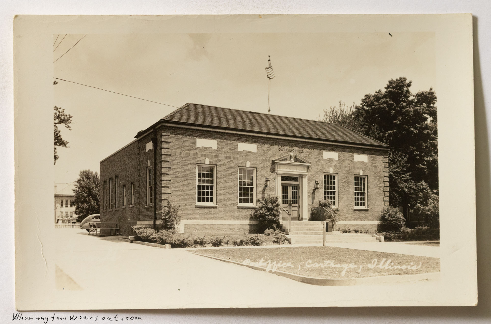 Front of postcard #1089: A real photo postcard showing the brick building of the Carthage, Illinois Post Office. The building has a flag flying f