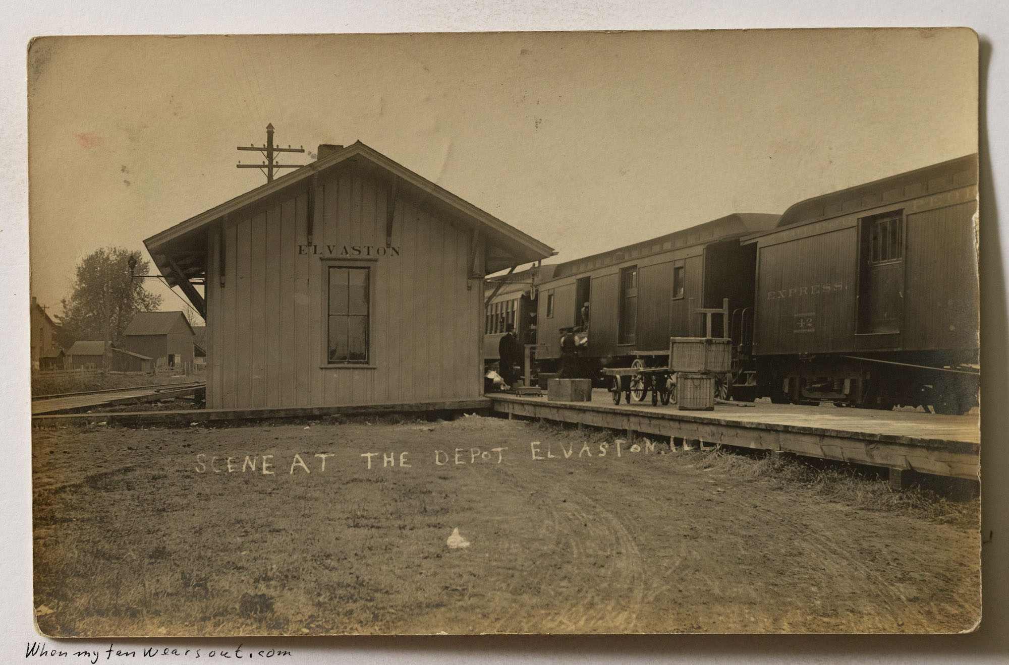 Front of postcard #1102: Sepia-toned photograph of the train depot in Elvaston, Illinois. The image shows a small wooden station building with "E